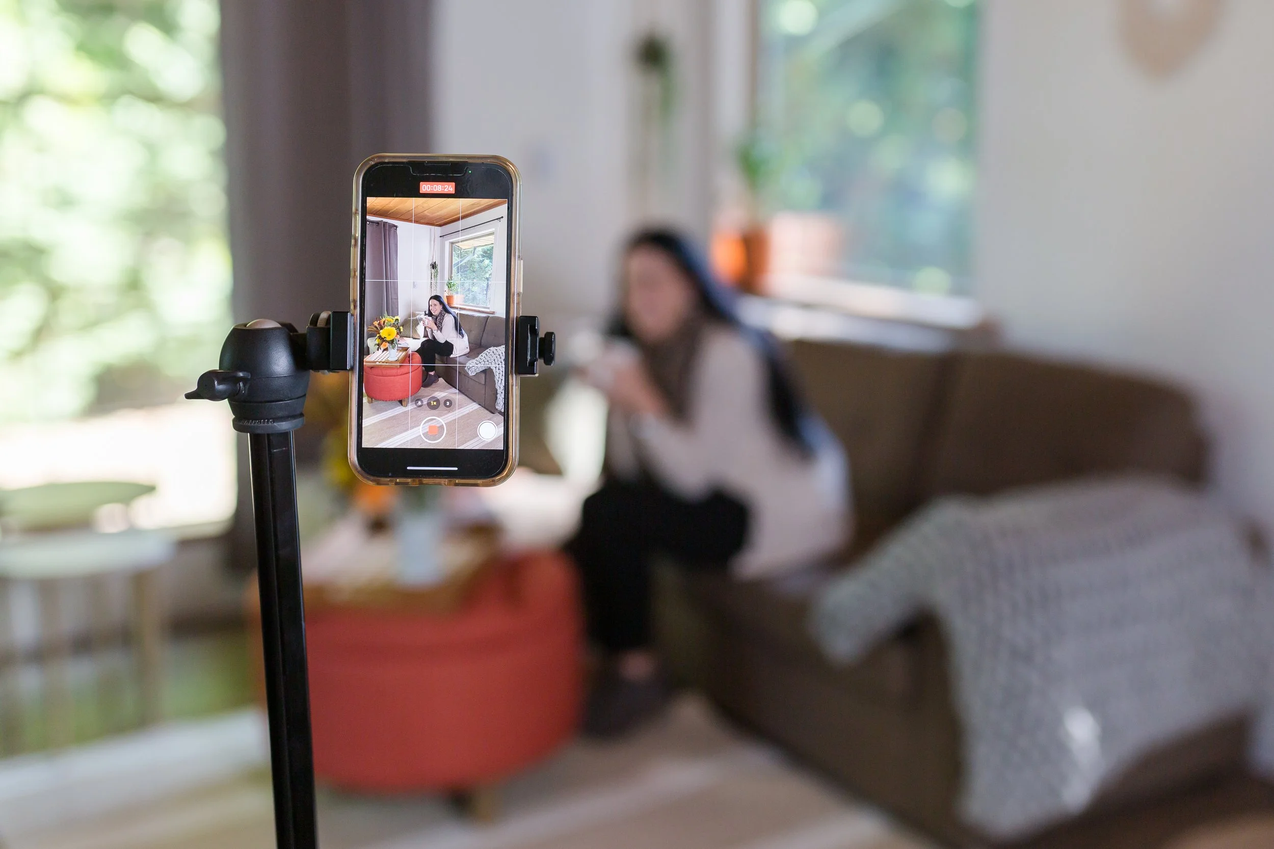 A smartphone mounted on a stand recording a woman sitting on a couch in a living room with a window in the background.