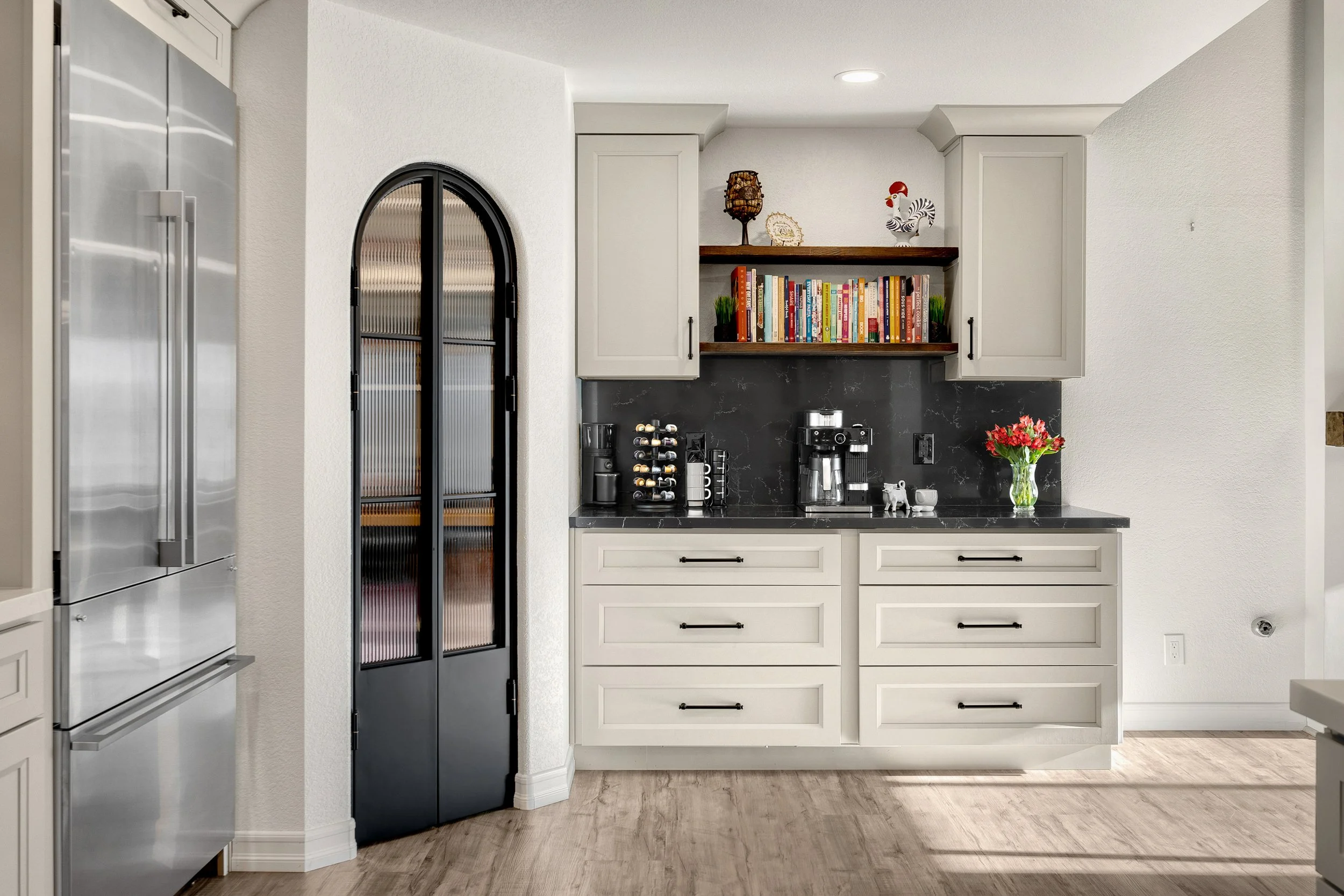 A modern kitchen with white cabinets, black countertop, coffee maker, espresso machine, wine bottles, and a bookshelf with books and decorative items on the wall.