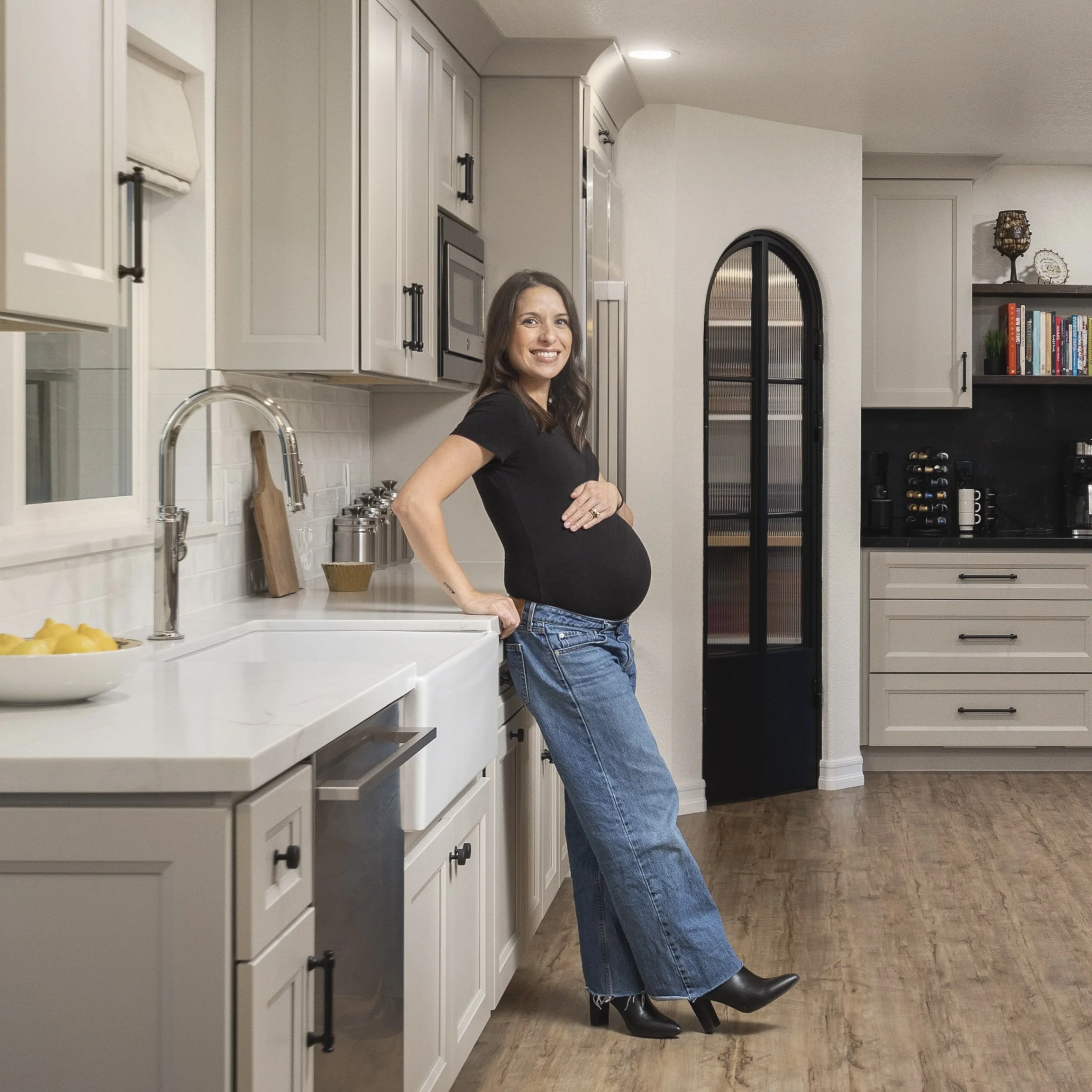 A pregnant woman with long brown hair, wearing a black t-shirt, jeans, and black high heels, leaning against a kitchen counter, smiling at the camera.