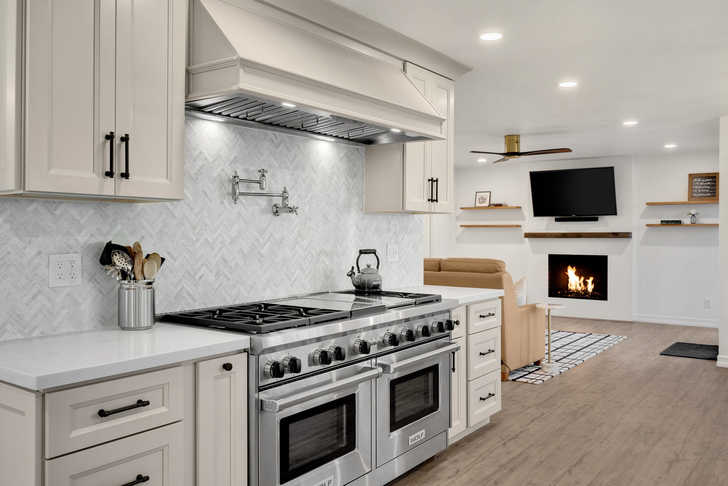 Modern kitchen with white cabinets, stainless steel stove, and a living room with a fireplace and TV in the background.