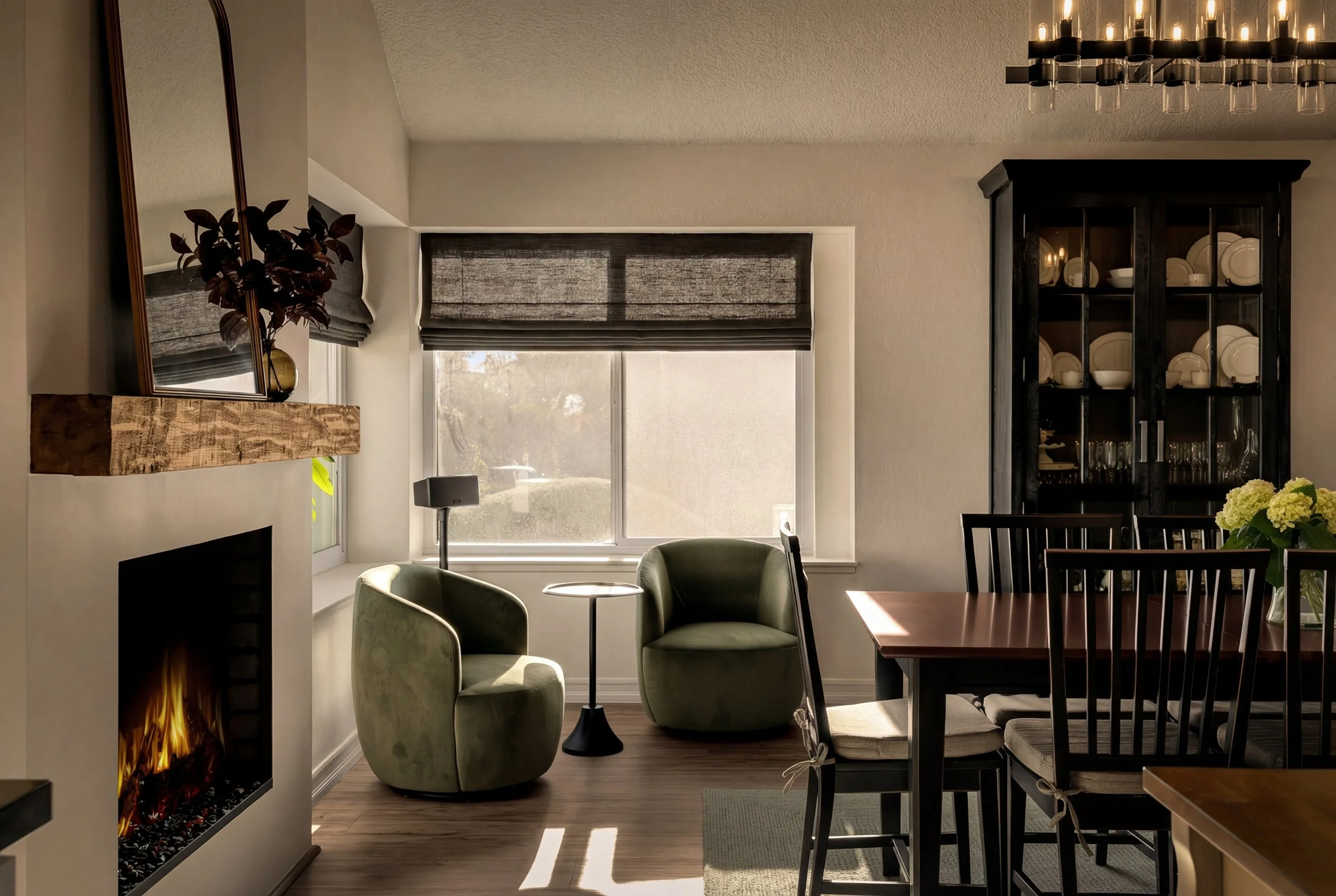 A cozy living room with a fireplace, two green armchairs, a small round table, and a large window with a dark shade. There's a black dining table with chairs and a black cabinet with plates and glassware, all illuminated by warm lighting.