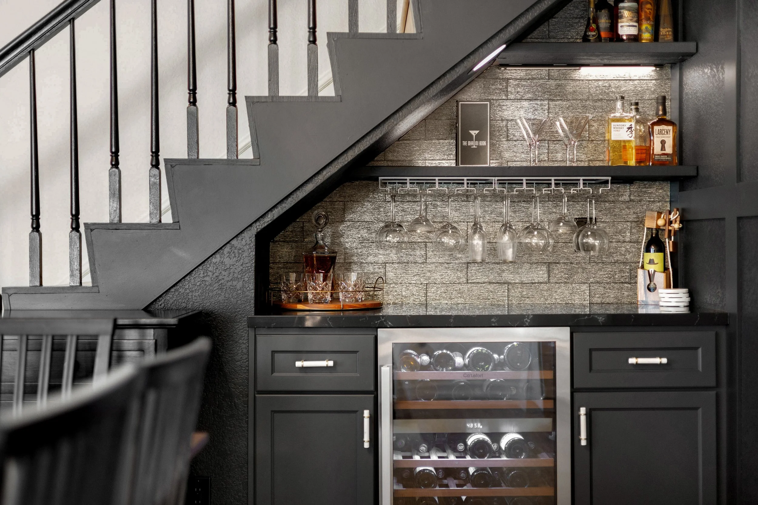 A black bar cabinet under a staircase with hanging wine glasses, liquor bottles, and a wine fridge.