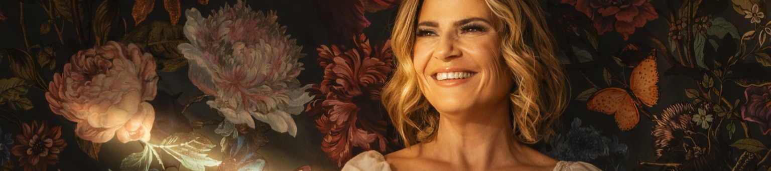 A smiling woman with shoulder-length wavy blonde hair against a dark floral background with pink and purple flowers and a butterfly.