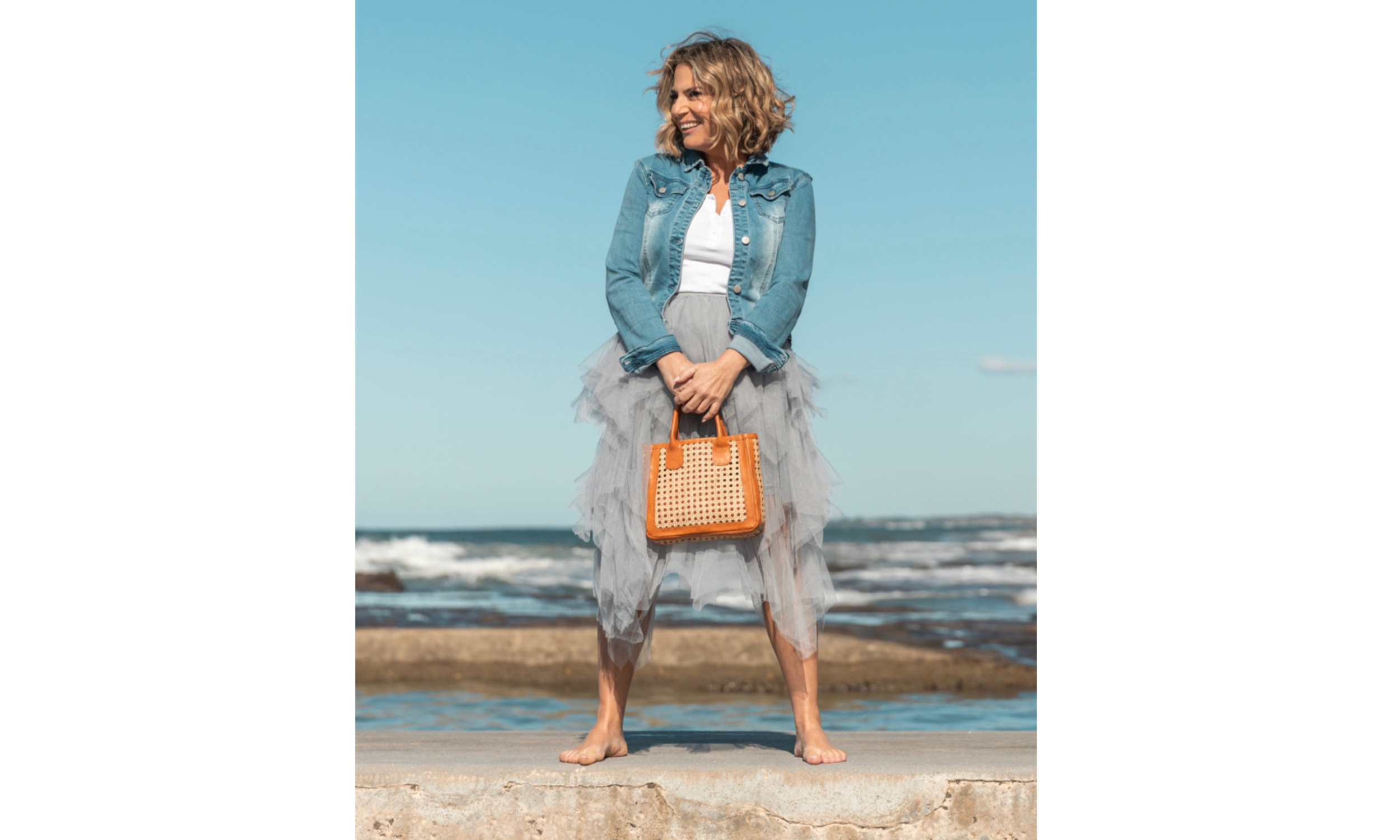 Woman standing barefoot on a beach with ocean in the background, wearing a denim jacket, white top, voluminous grey tulle skirt, and holding an orange woven handbag.