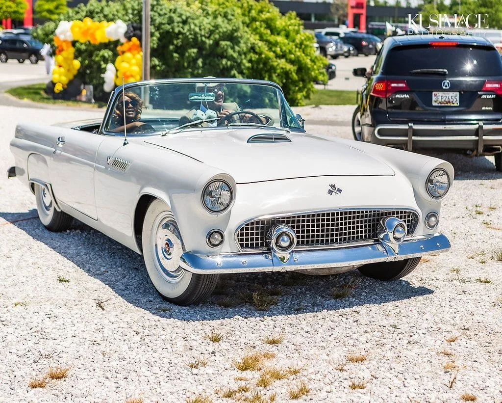 A vintage white convertible car parked outdoors with two smiling people inside, featuring a distinctive front grille and round headlights, with balloons and trees in the background.
