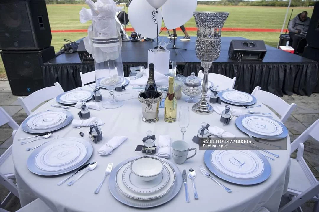 Elegant round dining table set outdoors with white tablecloth, white plates, silverware, napkins, and black place cards. The centerpiece includes champagne bottles, a large crystal candle holder, and white balloons. In the background, a stage with sound equipment and a grassy field are visible.