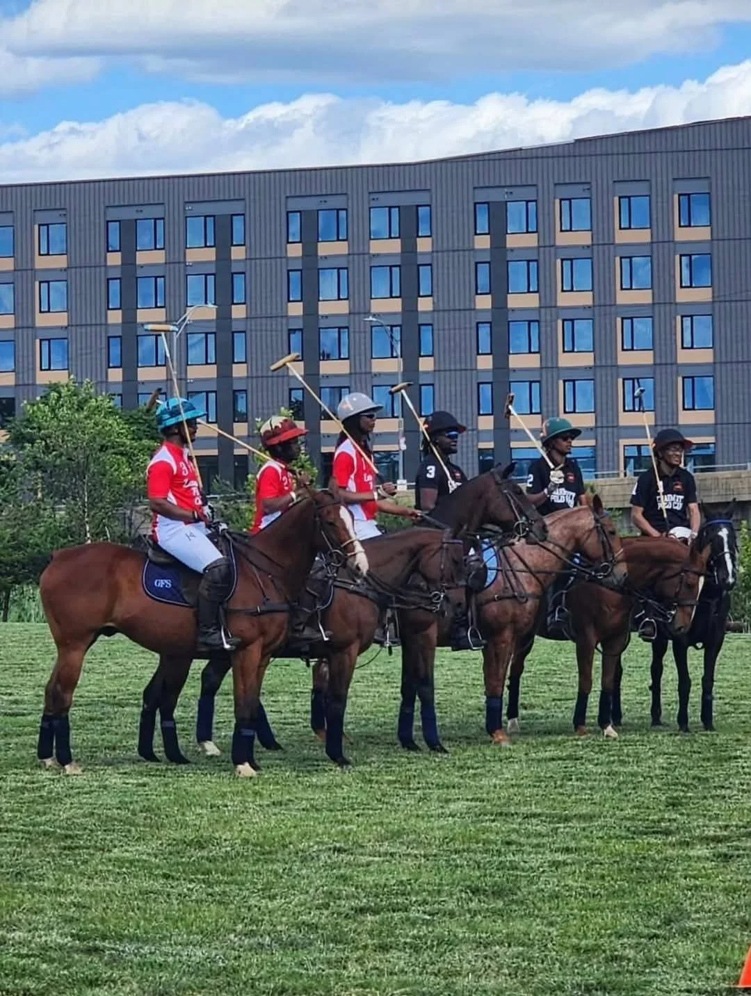 Six polo players on horseback standing on a grassy field, holding mallets, with a modern multi-story building in the background.