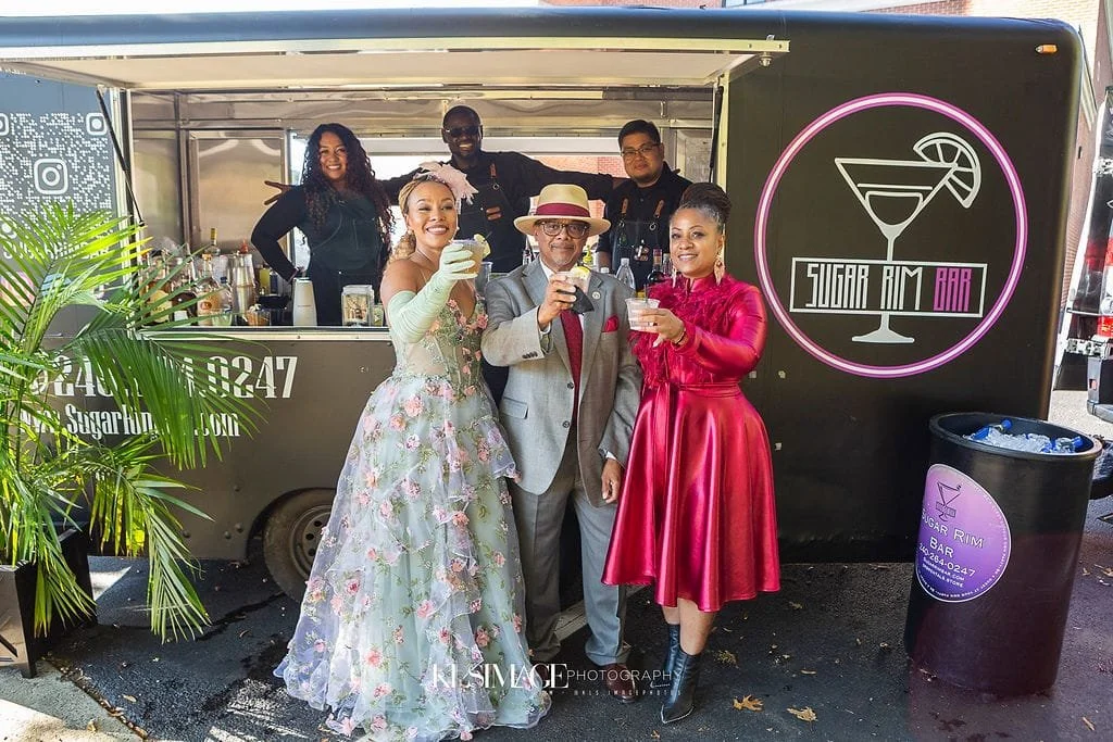 Group of people enjoying drinks in front of a black mobile bar called Sugar Rim Bar, with three staff behind the bar, and a decorative plant on the left side.