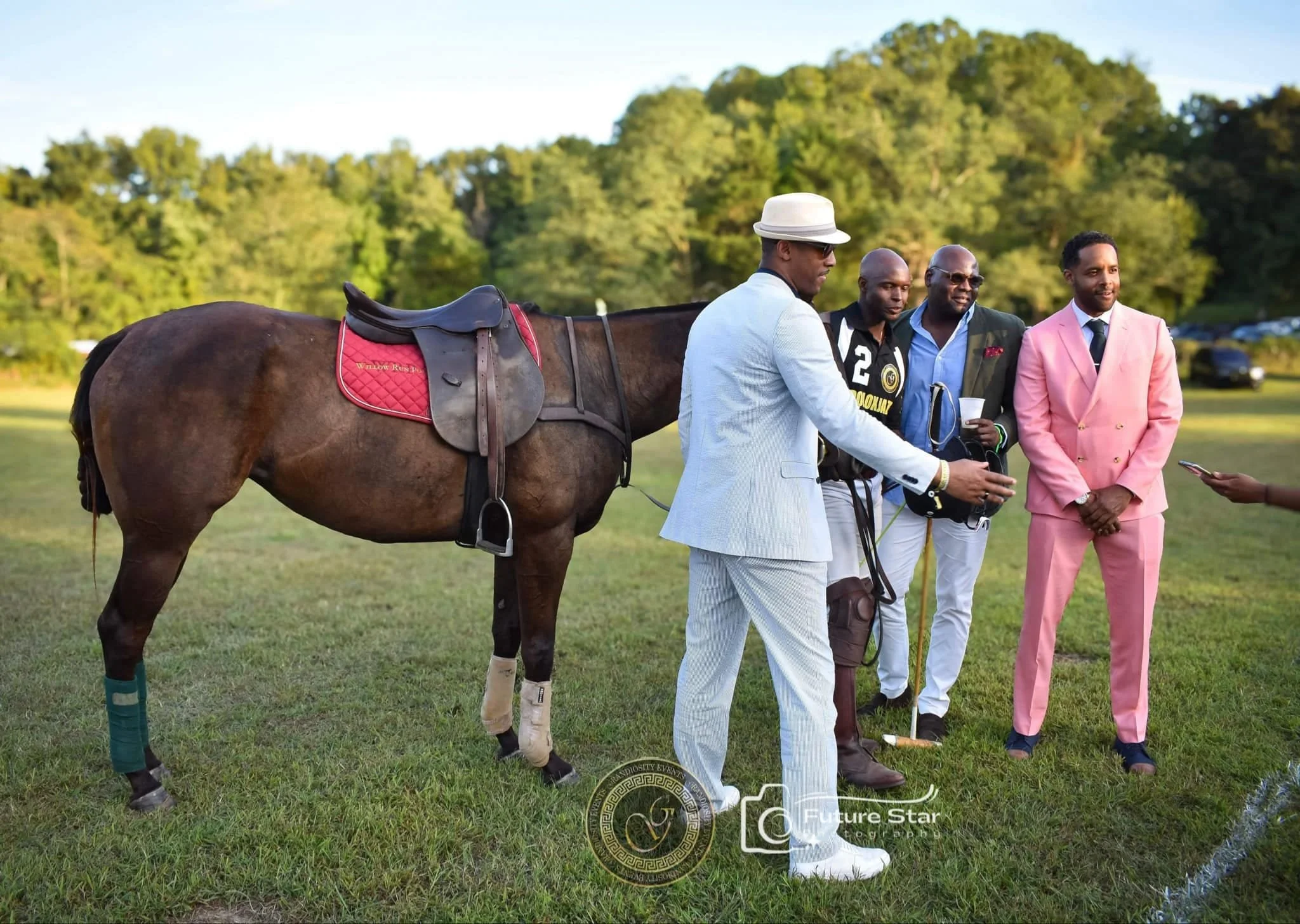 A group of five men standing on a grassy field near a brown horse with riding gear. The men are dressed in colorful suits, with one in pink, one in white, and others in darker colors. They are engaged in conversation, with some holding cups and a phone. The background shows trees and a blue sky, indicating a sunny day.