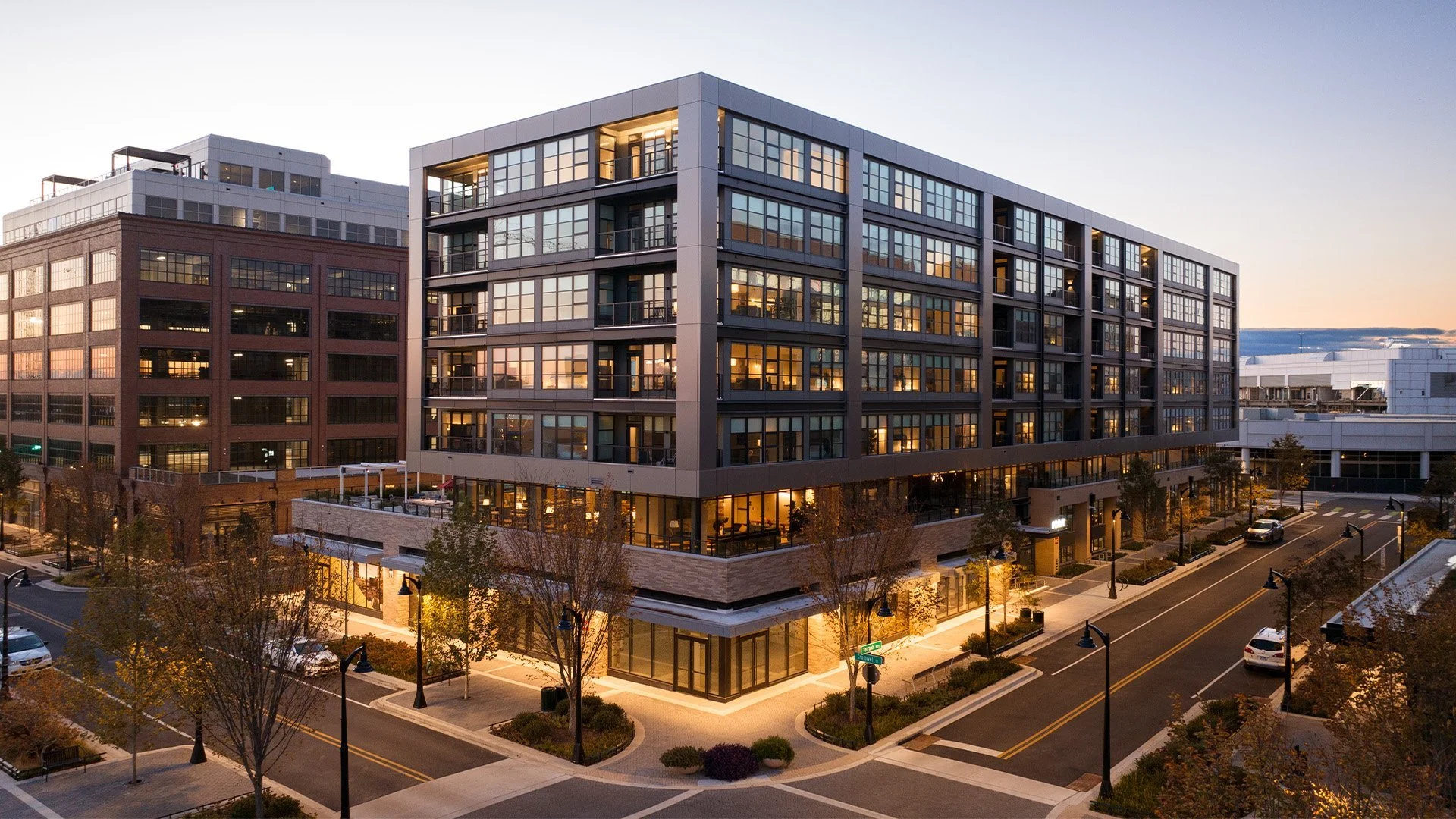 Modern multi-story apartment building with large glass windows, illuminated from inside, located at the corner of a well-lit street during dusk, with trees and parked cars visible.