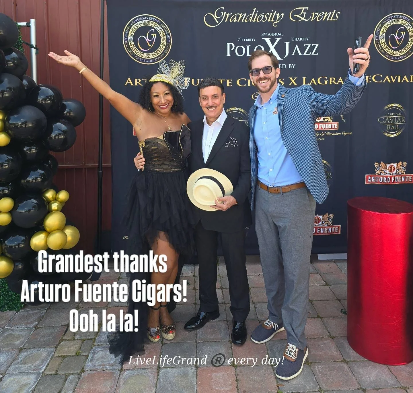 Three people pose together at a celebration event, standing in front of a black backdrop with gold and white text. The woman on the left wears a black strapless dress with a gold fascinator hat, the man in the middle wears a black suit holding a straw hat, and the man on the right wears a blue blazer and gray trousers. They are smiling and waving at the camera. There are balloon decorations on the left and a red pedestal on the right. The overlay text reads 'Grandest thanks Arturo Fuente Cigars! Ooh la la! LiveLifeGrand every day!'.