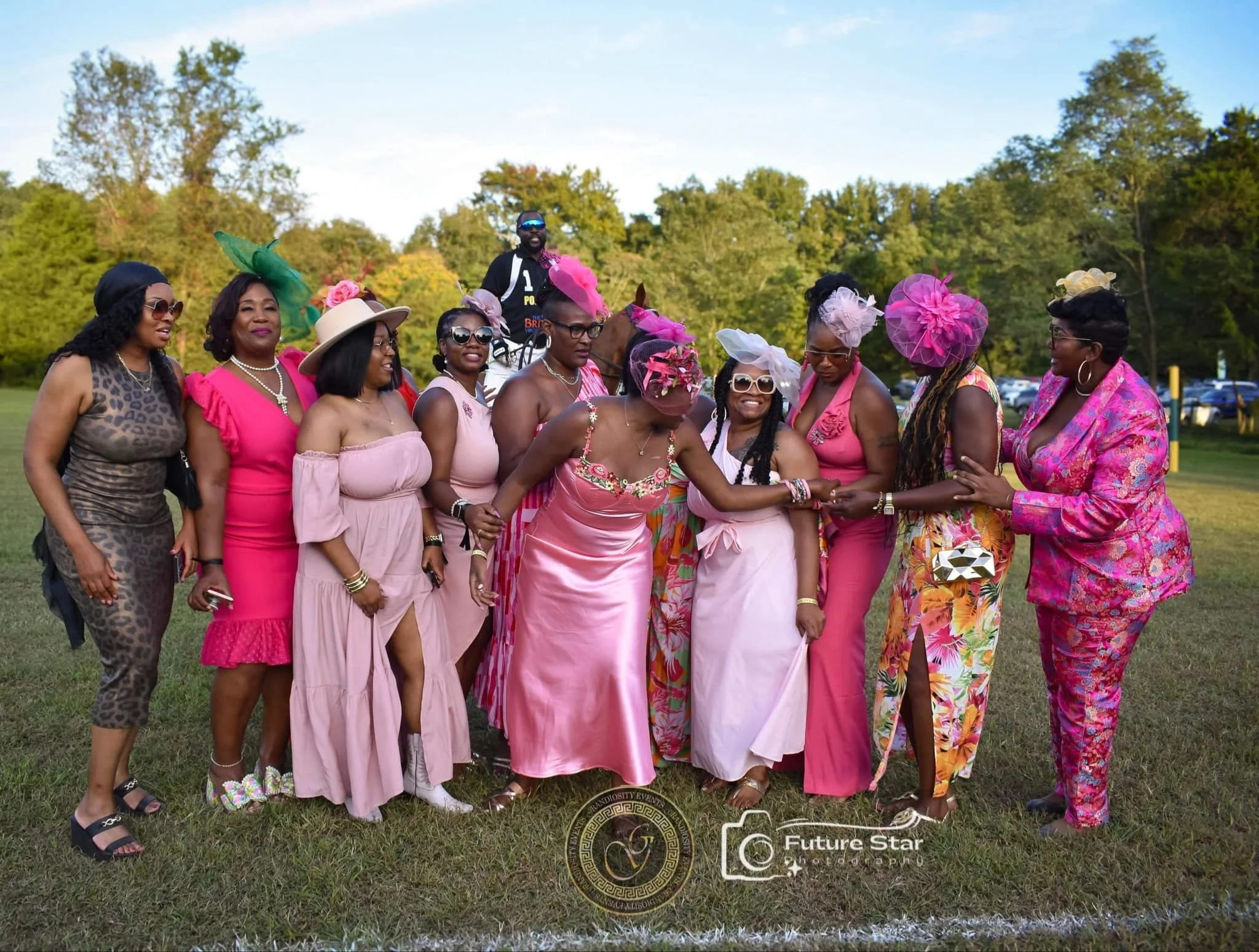 A group of women dressed in colorful pink and floral dresses with hats and fascinators, holding hands in a circle outdoors on grass, with trees and a blue sky in the background.