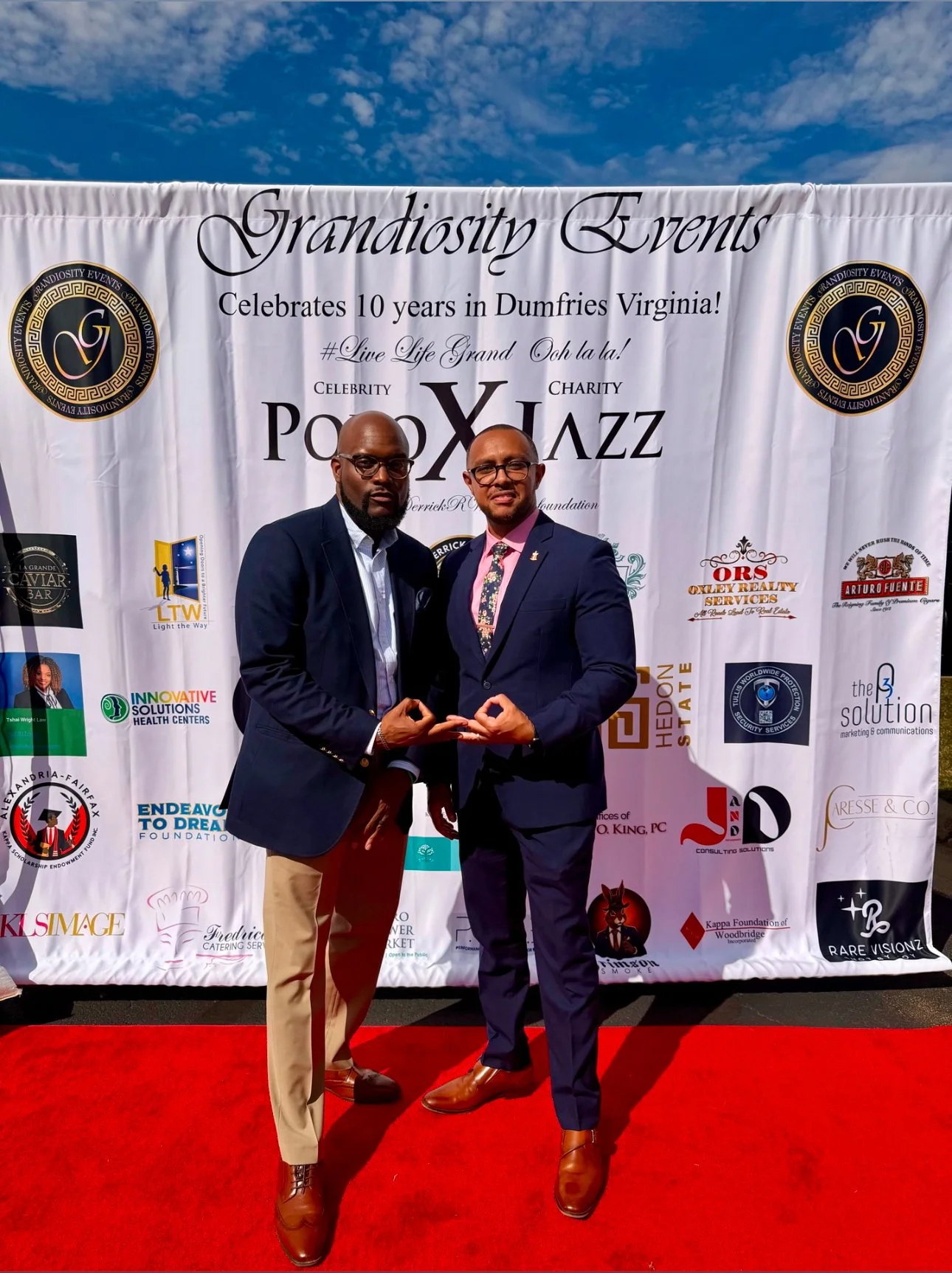 Two men in formal suits posing on a red carpet in front of a white promotional banner celebrating 10 years of Grandiosity Events in Dumfries, Virginia. They are standing close together, making a hand gesture, with the banner displaying various logos and text about the event.