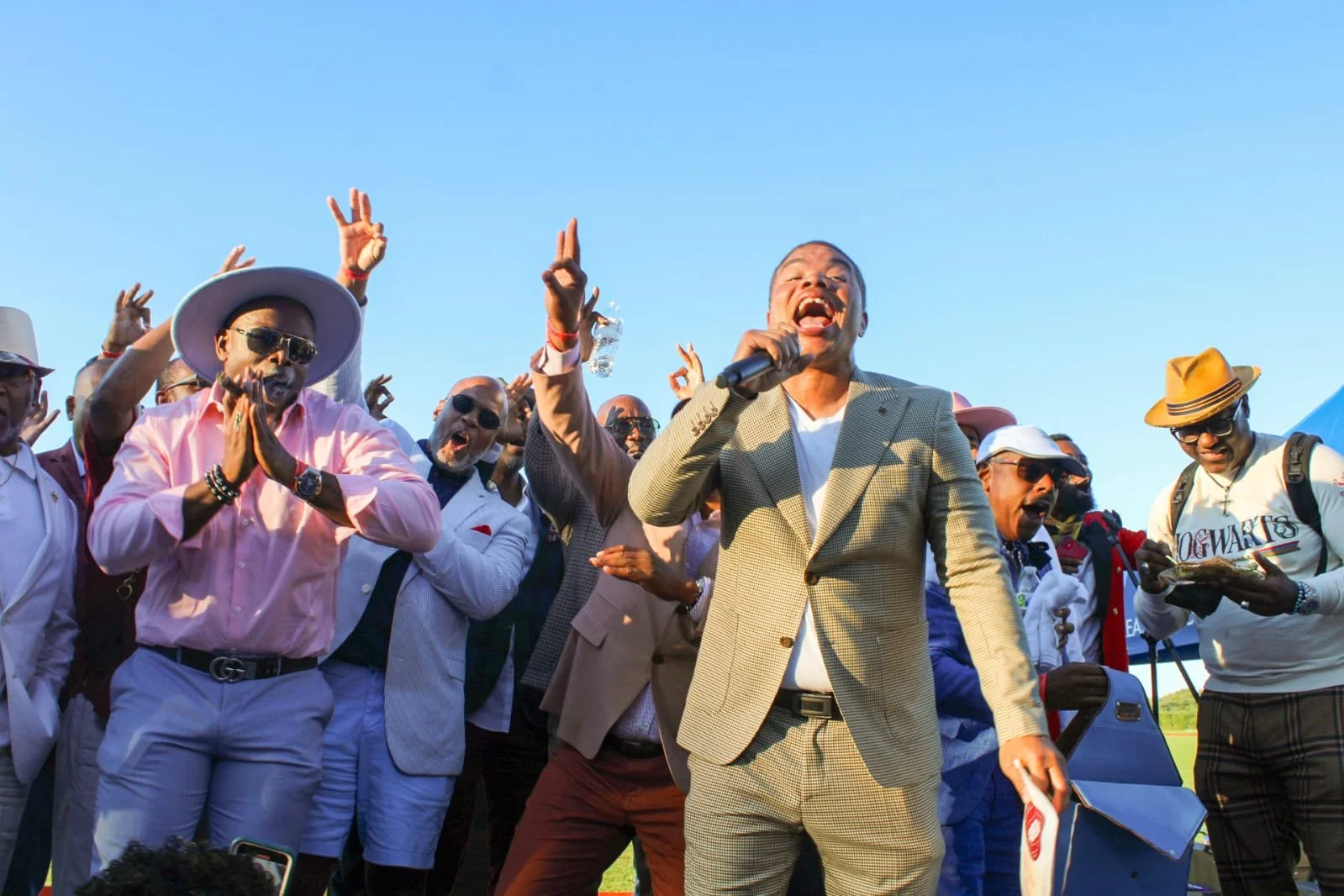 A group of diverse people outdoors, some wearing sunglasses and hats, celebrating with a man in a beige blazer speaking into a microphone under a clear blue sky.