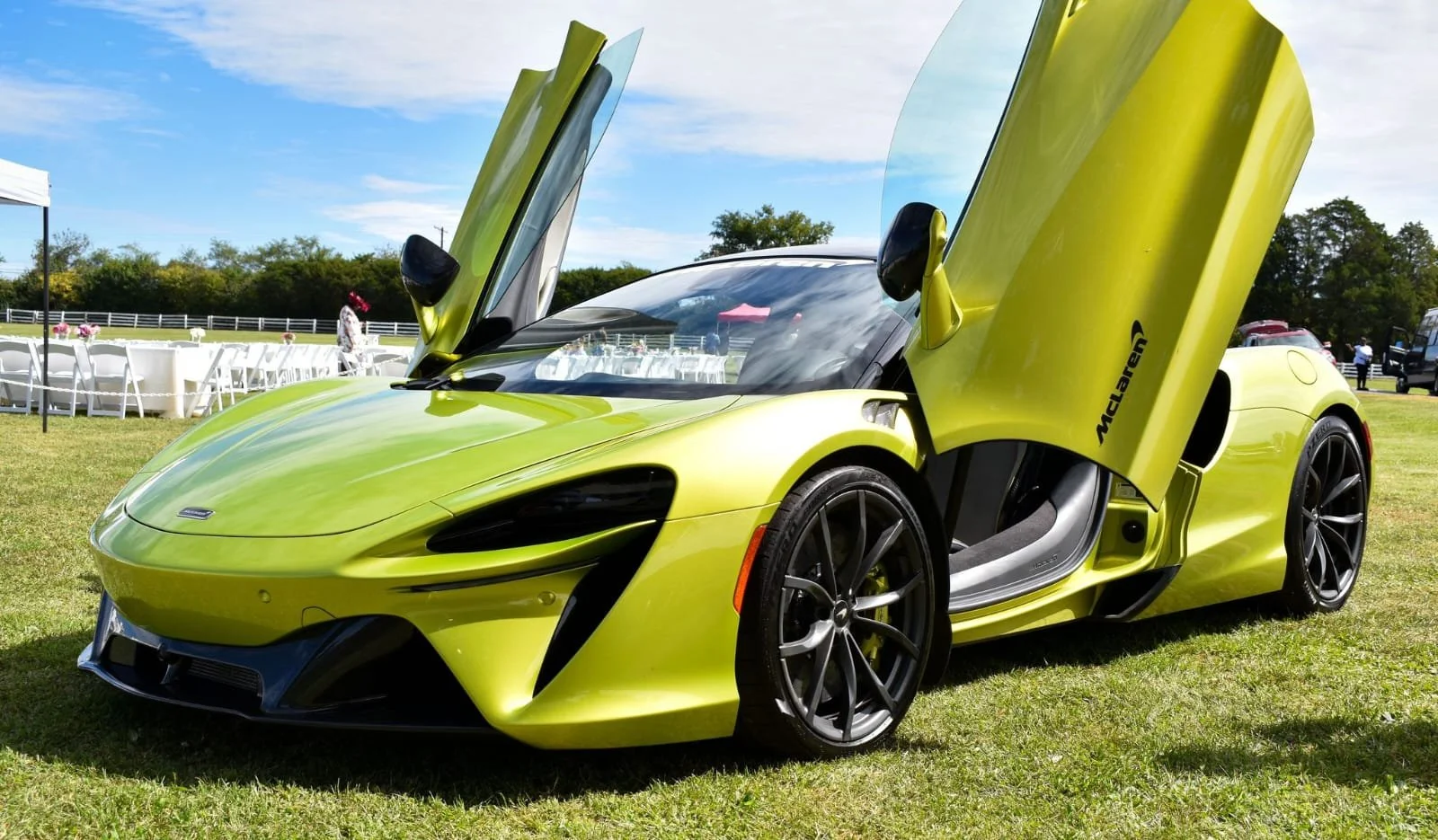A bright yellow-green McLaren sports car with its gullwing doors open, parked on grass at an outdoor event on a sunny day.