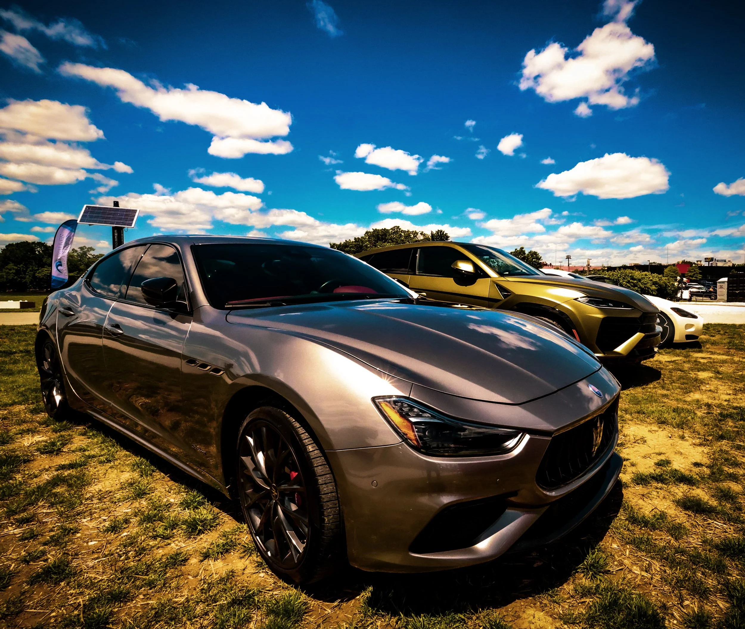 A lineup of luxury cars parked on grass under a blue sky with white clouds.