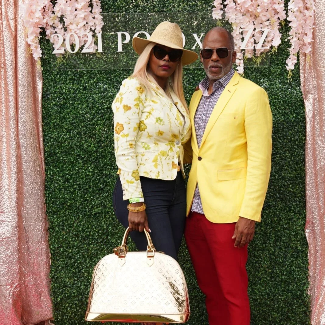 Two stylish adults, a woman and a man, posing outdoors in front of a decorative hedge backdrop with pink flowers and the year "2021" displayed. The woman wears a floral blazer, dark pants, sunglasses, a wide-brimmed hat, and carries a large white handbag. The man wears a yellow blazer, patterned shirt, red pants, and sunglasses.