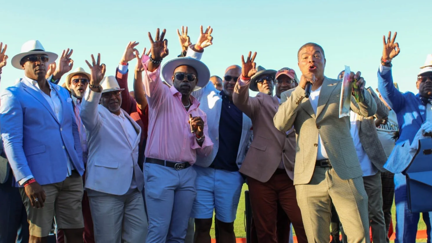 A group of men dressed in colorful suits and hats, some wearing sunglasses, gathered outdoors under a clear sky, raising their hands and holding a microphone, appearing to participate in a public event or celebration.