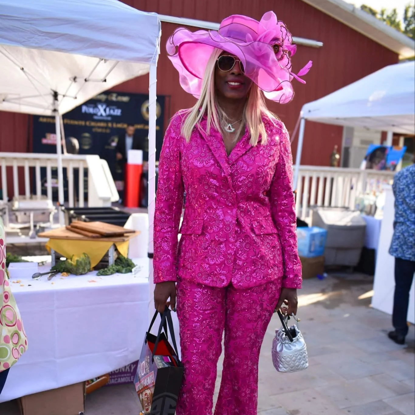 Woman wearing a bright pink lace suit, large pink hat with netting, sunglasses, and blonde hair, holding a small white quilted bag and a shopping bag, standing outdoors at an event with tents and tables.