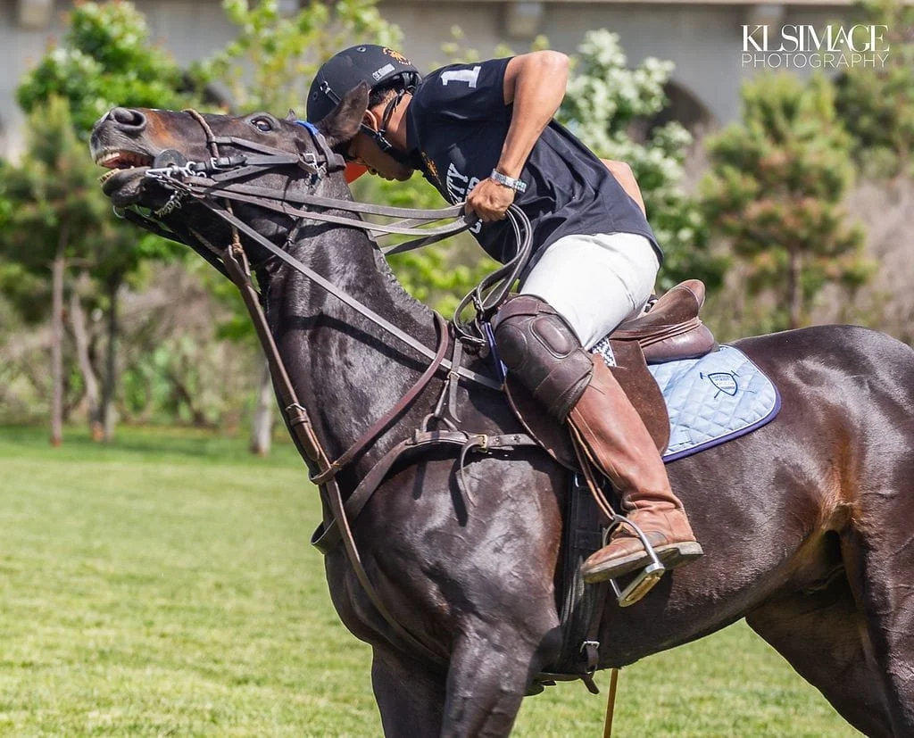 A jockey wearing a black helmet and shirt riding a dark brown horse with its mouth open during a race on a grassy field.