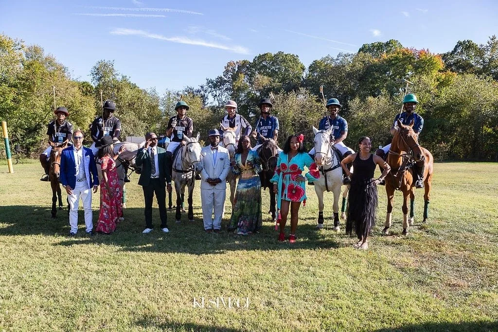 A group of people, some on horseback and others standing on the grass, during daytime with a backdrop of trees and a clear sky.
