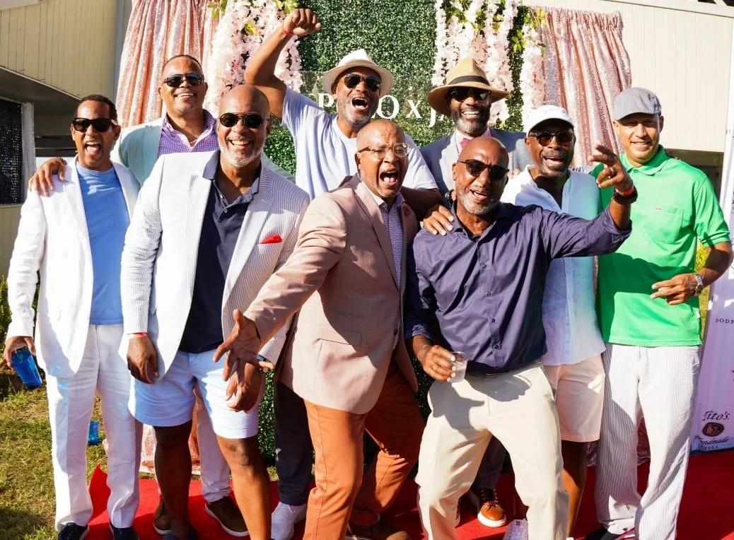 Group of nine men outdoors celebrating at a social event, with a green and pink backdrop and sunlight, wearing casual and semi-formal attire, some in sunglasses and hats, posing and smiling.
