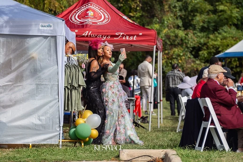 Two women in fancy gowns taking a selfie at an outdoor event under a red tent that reads "Always First." There are tables with people dining and a line of balloons on the ground.