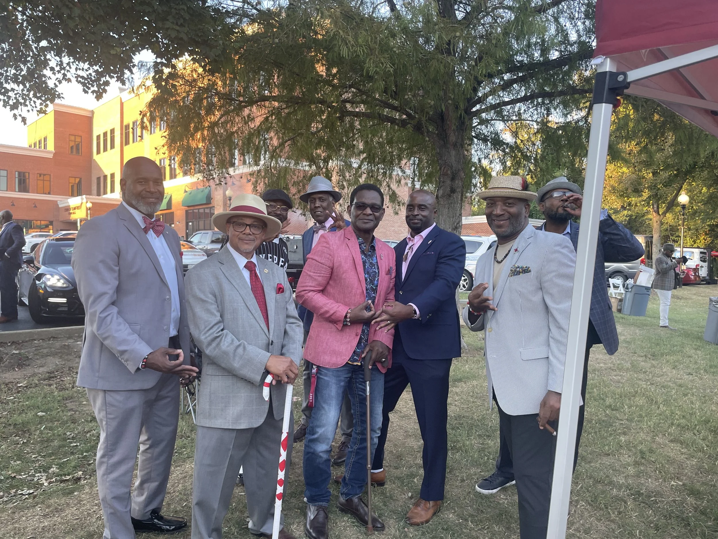 A group of seven men dressed in suits and casual attire, standing outdoors under a tree, at an event or gathering in a city park or plaza, with parked cars and a building in the background.