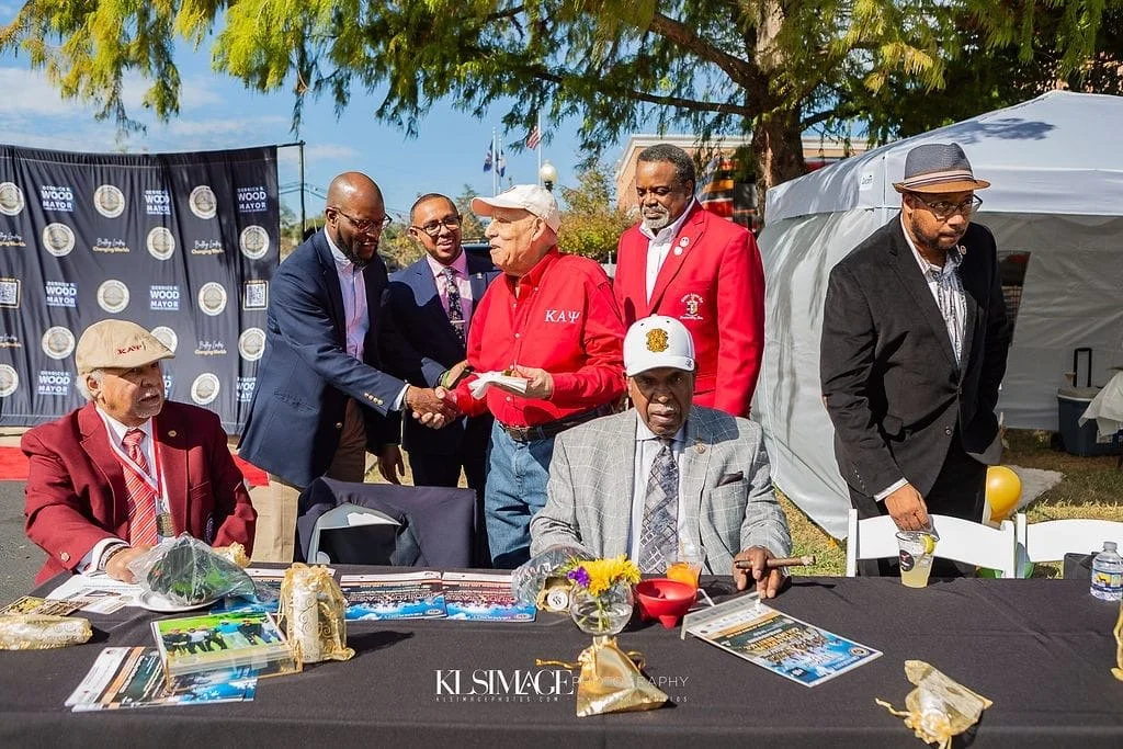 A group of men at an outdoor event, with some shaking hands. They are wearing formal and semi-formal attire, and there is a table with magazines, flowers, and decorations in front of them. The background includes a banner, trees, and tents, indicating a ceremony or celebration.