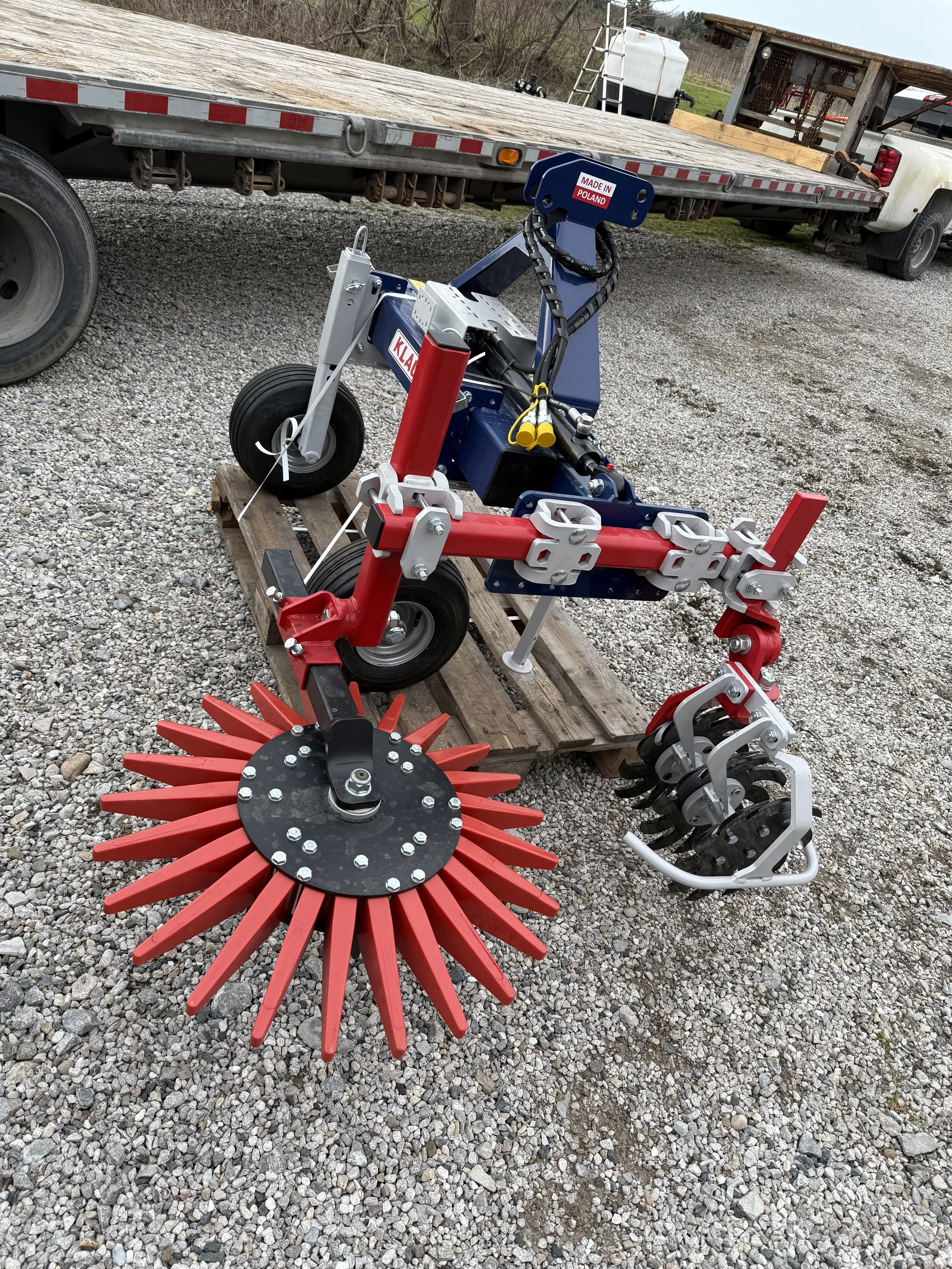 A  piece of agricultural machinery with wheels, call Klaudia sitting on a wooden pallet outdoors on gravel. It has a large, circular, red brush at the front, and multiple black and gray tines at the back, and is labeled "Made in Poland."