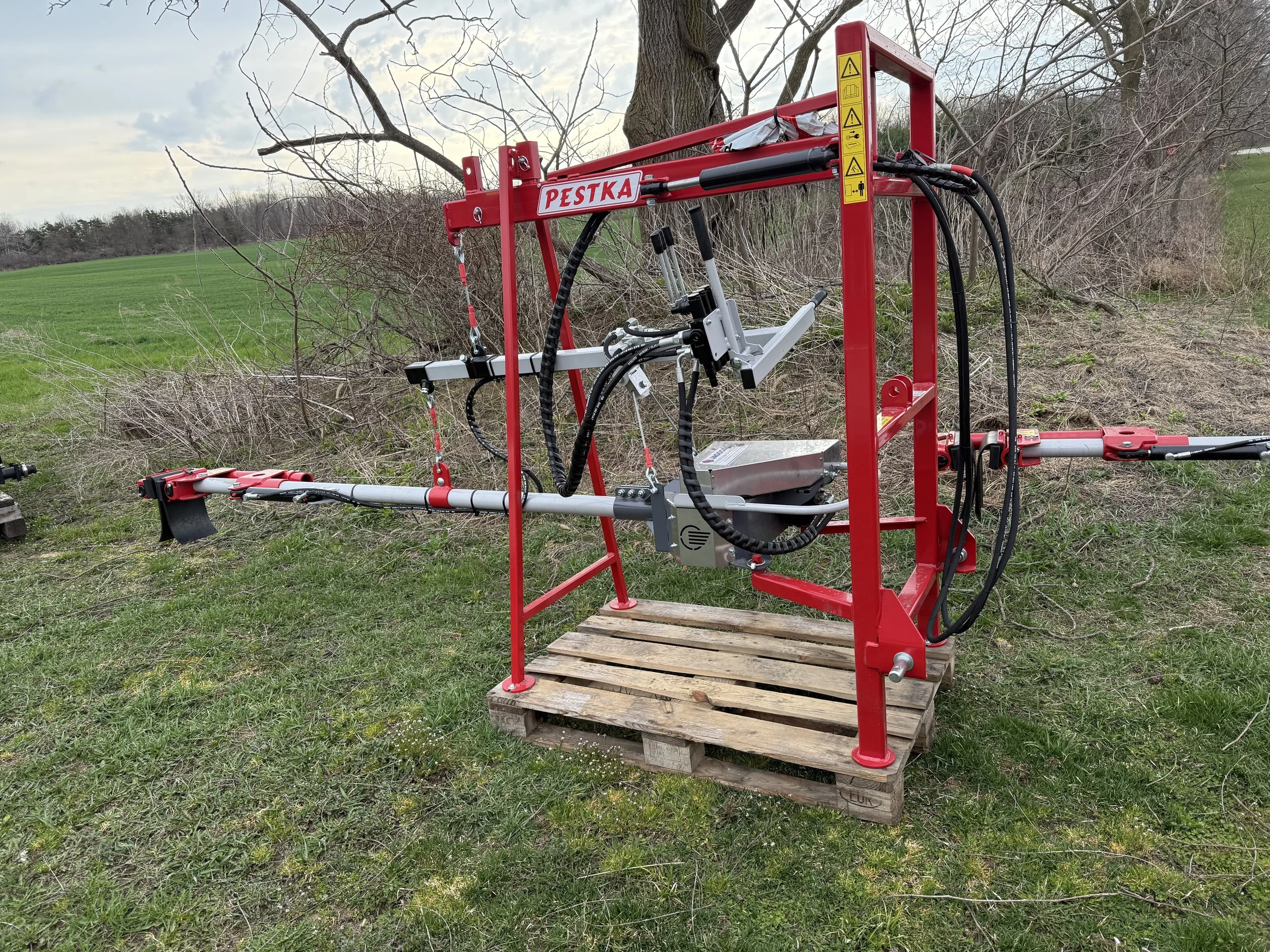 A red  Jagoda Tree shaker called Pestka , an agricultural tool with hydraulic components, sitting on a wooden pallet on grass in an open field.