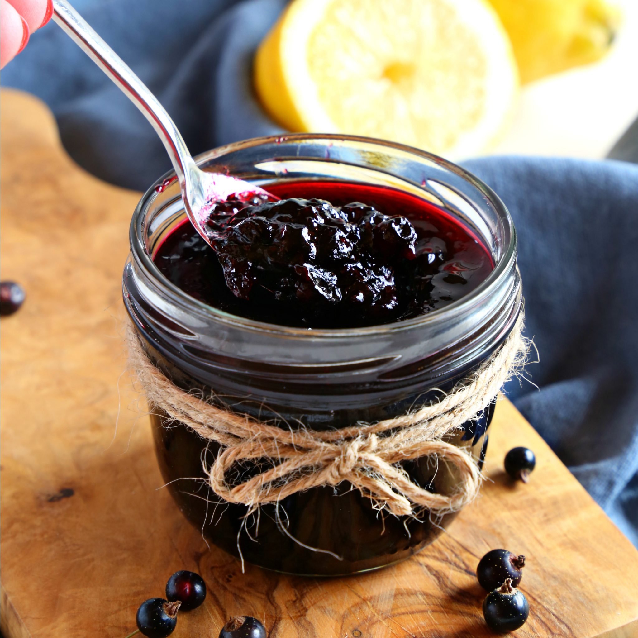 A jar of dark blackcurrant jam with a spoon being dipped into it, placed on a wooden surface with lemon slices in the background.