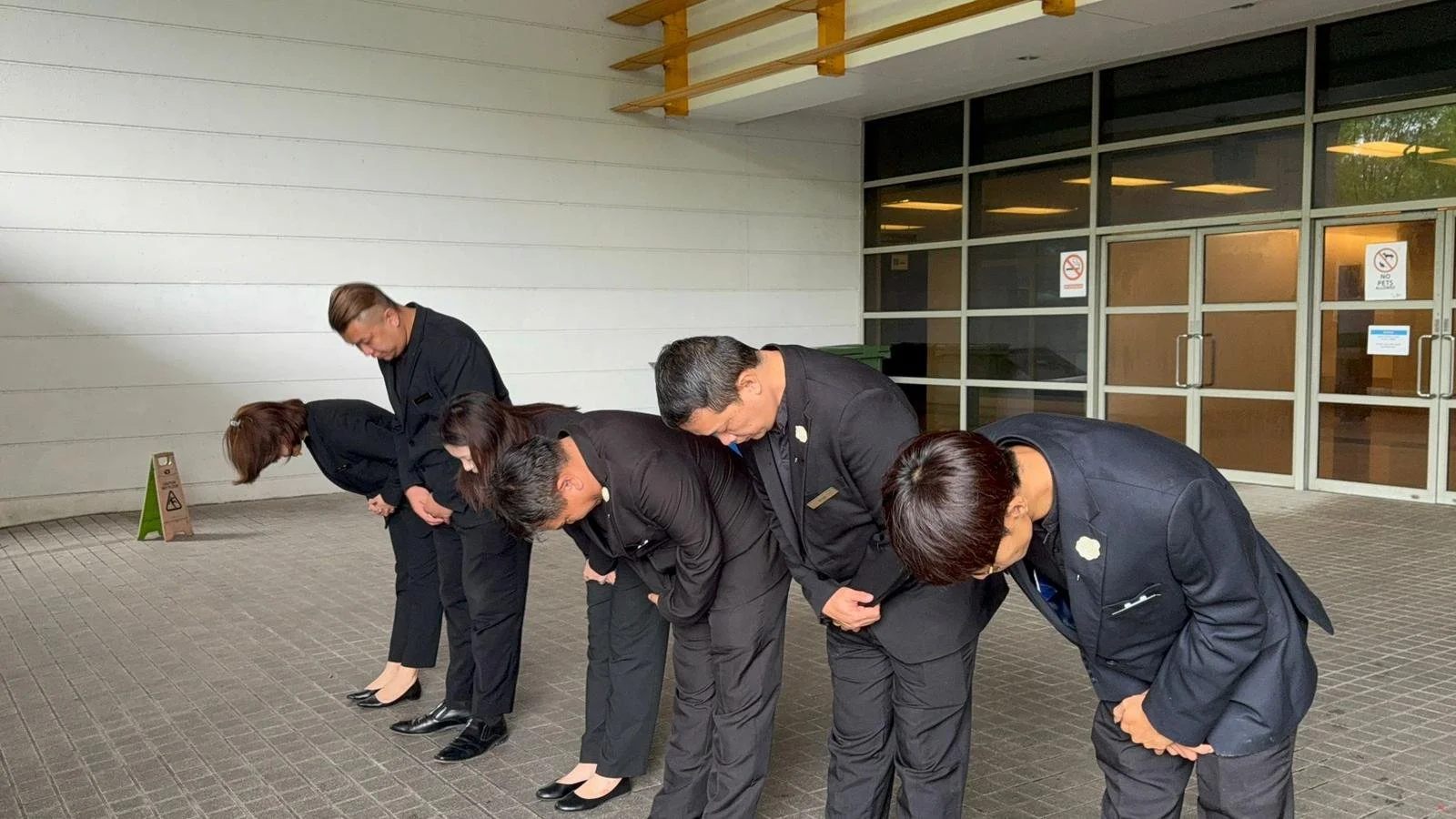 Five people in business attire bowing in front of a building entrance.