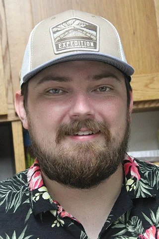 A man with a beard and mustache wearing a beige trucker hat with a mountain expedition patch, and a black Hawaiian shirt with pink and yellow floral patterns, standing in front of wooden cabinets.