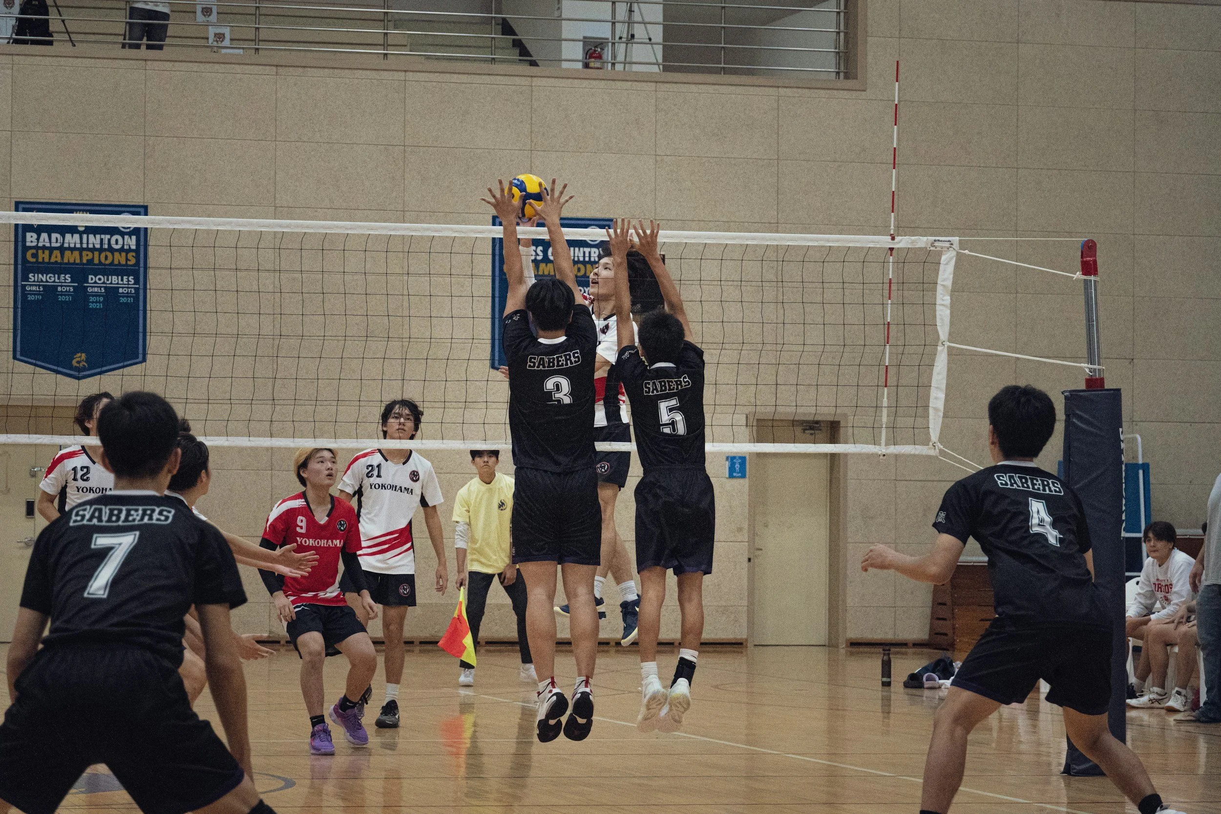 Players from two volleyball teams in action during a match in an indoor gymnasium, with two players jumping to block the ball at the net, and other players ready for the play.