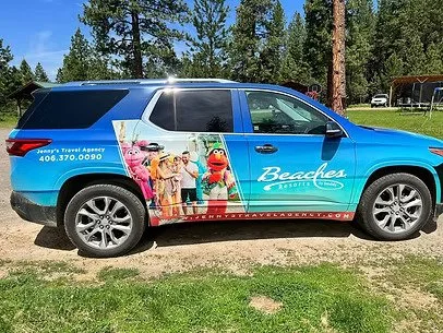 A blue SUV with advertising for Beaches Resort featuring a cartoon character and children, parked on grassy ground with trees in the background.