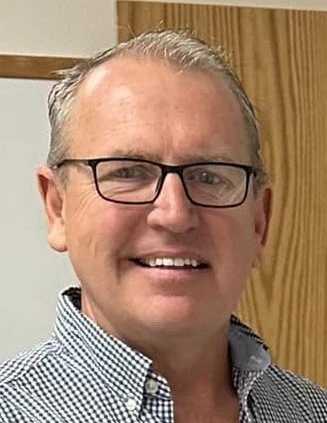 Portrait of a middle-aged man with short gray hair wearing a blue and white checkered button-up shirt against a plain gray background.