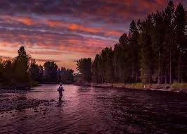 A person fishing in a river during sunset with colorful sky and trees along the riverbanks.