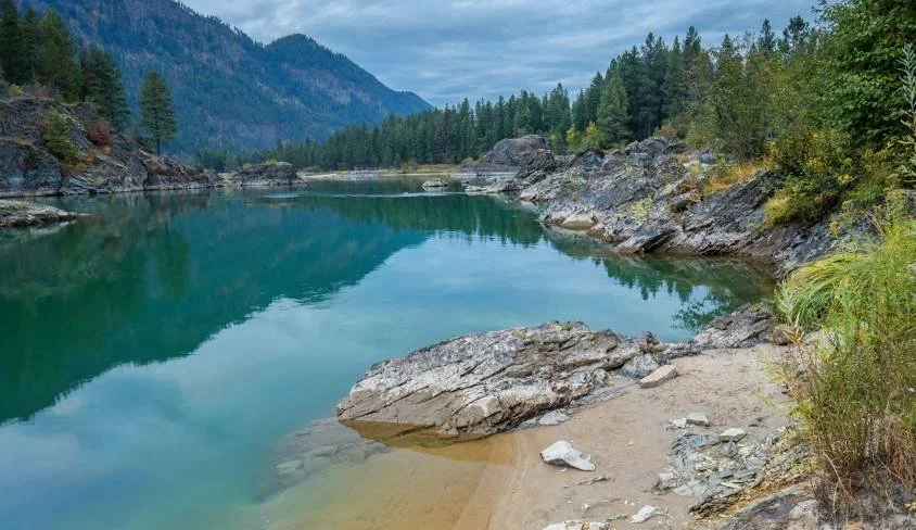 A calm river with clear, turquoise water flowing between rocky banks surrounded by green trees and mountains in the distance under a cloudy sky.