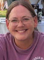 A smiling woman with glasses and brown hair, wearing a purple shirt, outdoors near a vehicle.