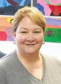 Smiling woman with light brown hair and gray shirt standing in front of colorful painted background.