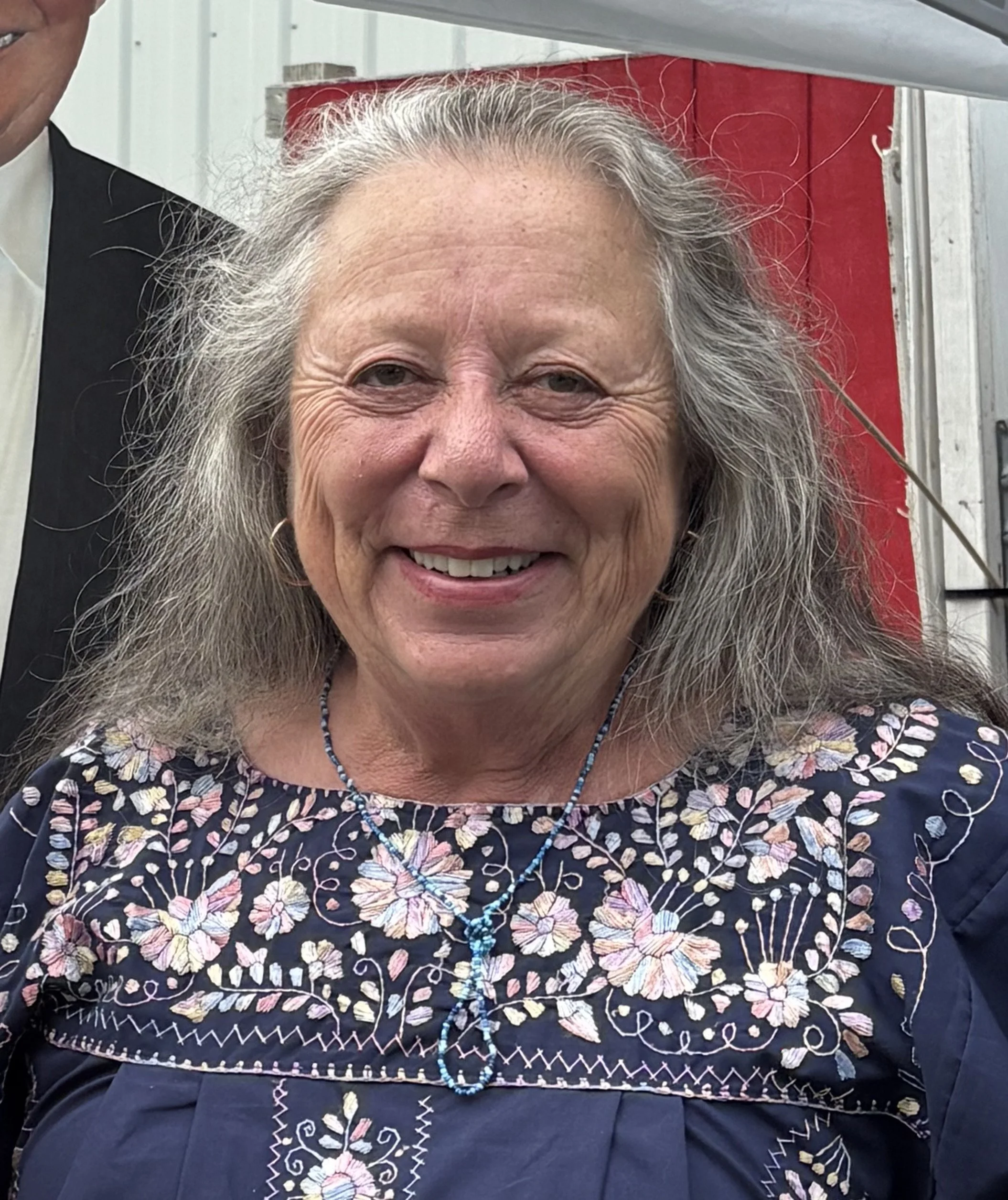 Close-up of an elderly woman with gray hair, smiling, wearing a dark floral embroidered blouse and a beaded necklace.