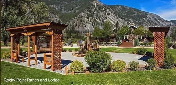 A landscaped outdoor area with two wooden gazebos, bushes, and trees, with mountains and lodge buildings in the background, labeled "Rocky Point Ranch and Lodge."