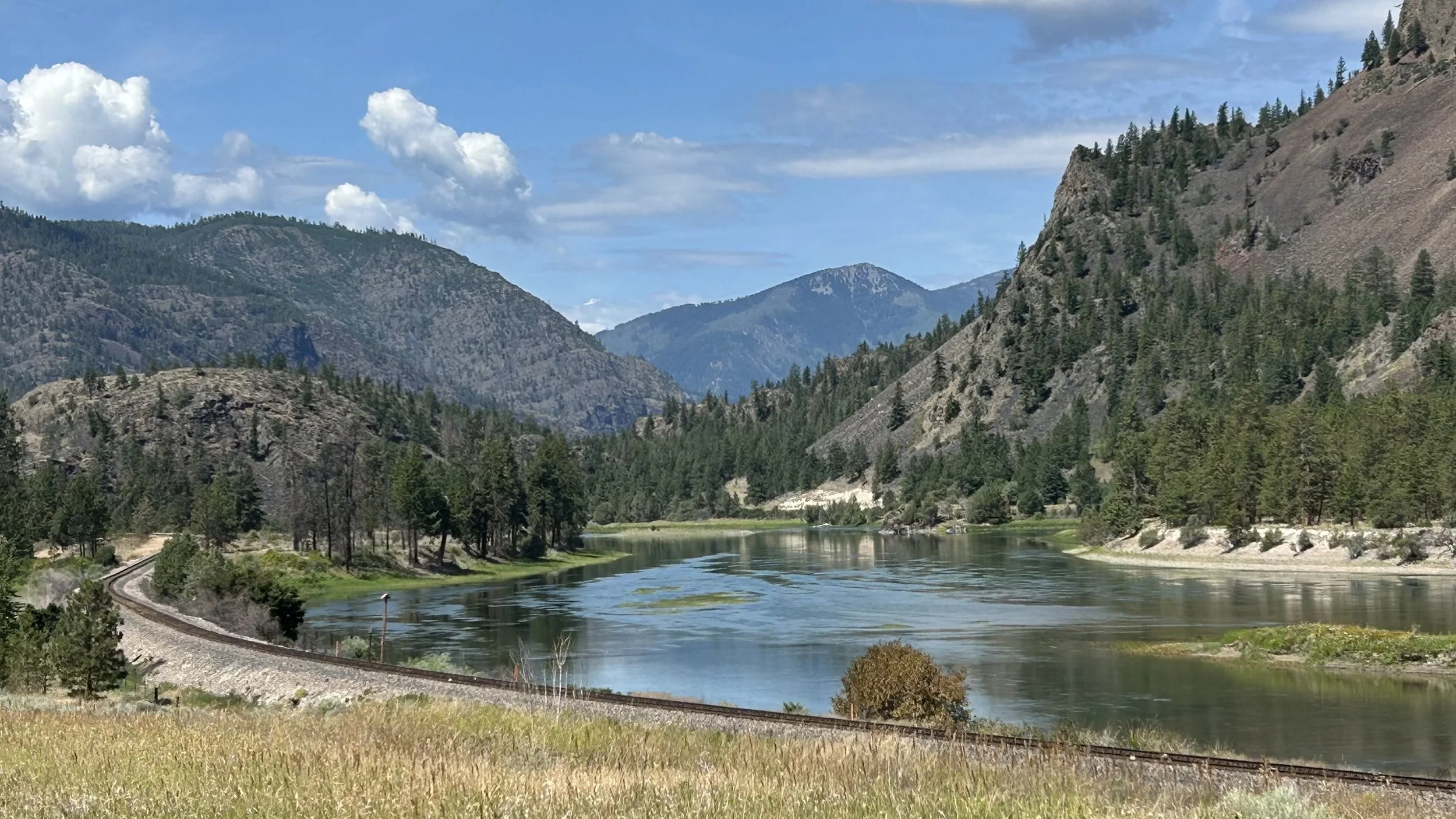 Scenic view of a mountain valley with a river, surrounded by forested hills and mountains under partly cloudy sky.