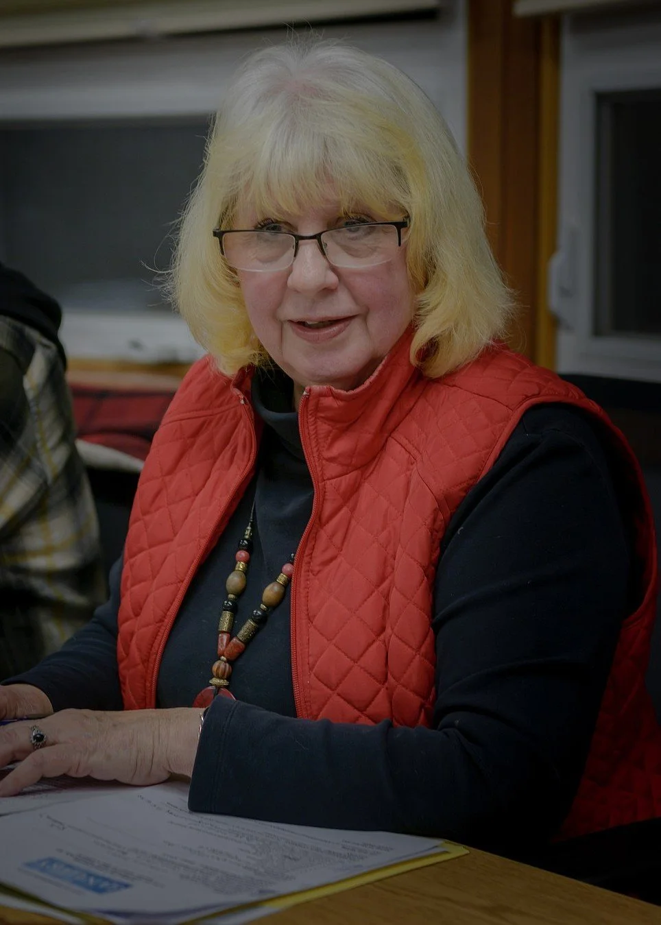 A woman with blonde hair, glasses, and a red quilted vest sitting at a table with papers in front of her, smiling.