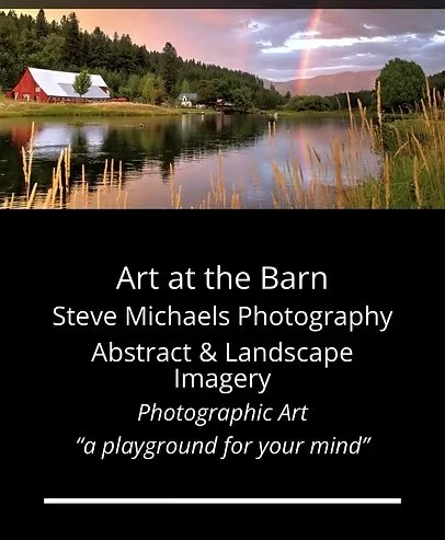 A landscape scene featuring a lake with tall grasses in the foreground, a red barn with a white roof on the left, dense trees in the background, a rainbow arching across the sky, and a partly cloudy sky at sunset.