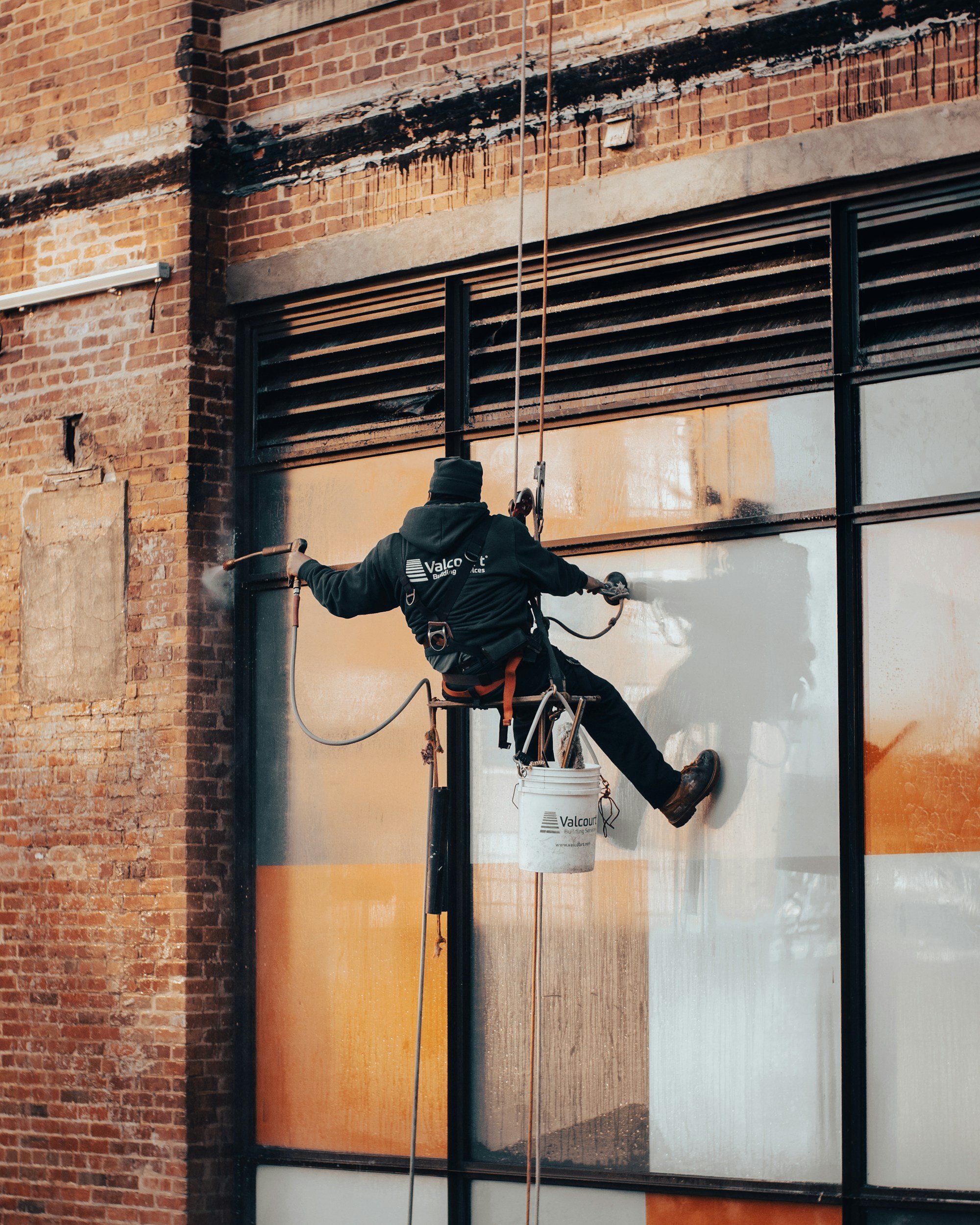 Worker cleaning large glass window on a brick building façade with a power washer, wearing safety harness and bucket.