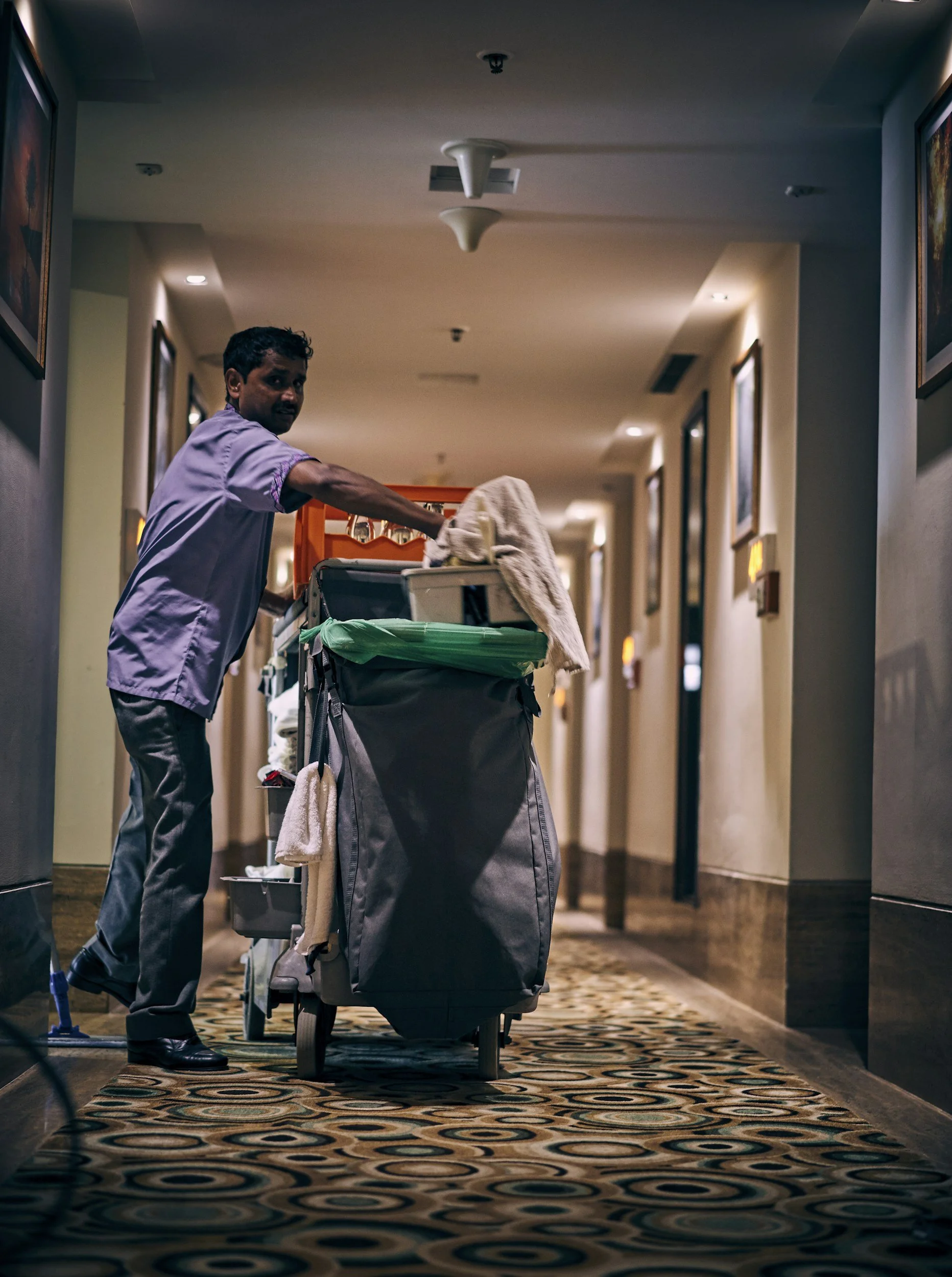 A hotel staff member pushing a cleaning cart down a hotel corridor.
