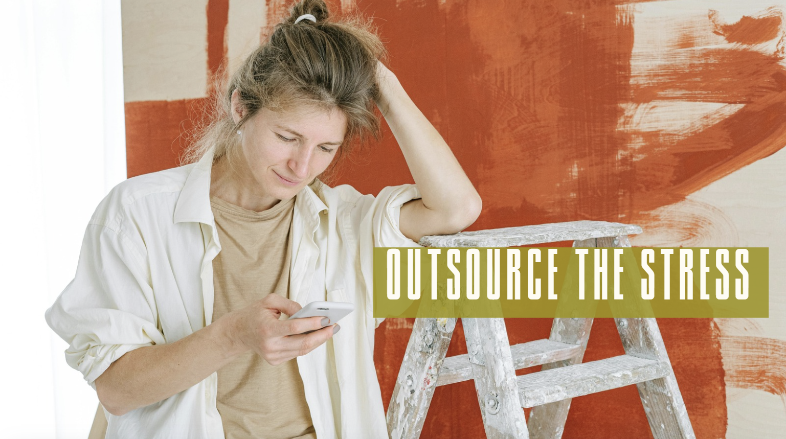 A woman in casual clothing looking at her phone with a puzzled or stressed expression, standing next to a wooden ladder against an orange painted wall with paint strokes.