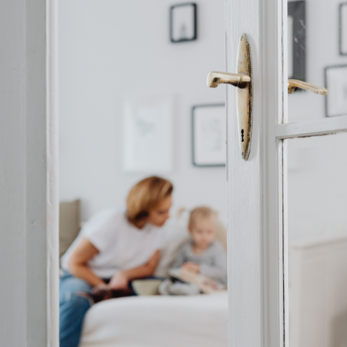 A white door is slightly open, showing a woman and a child sitting on a bed in a bedroom, with picture frames hanging on the wall behind them.